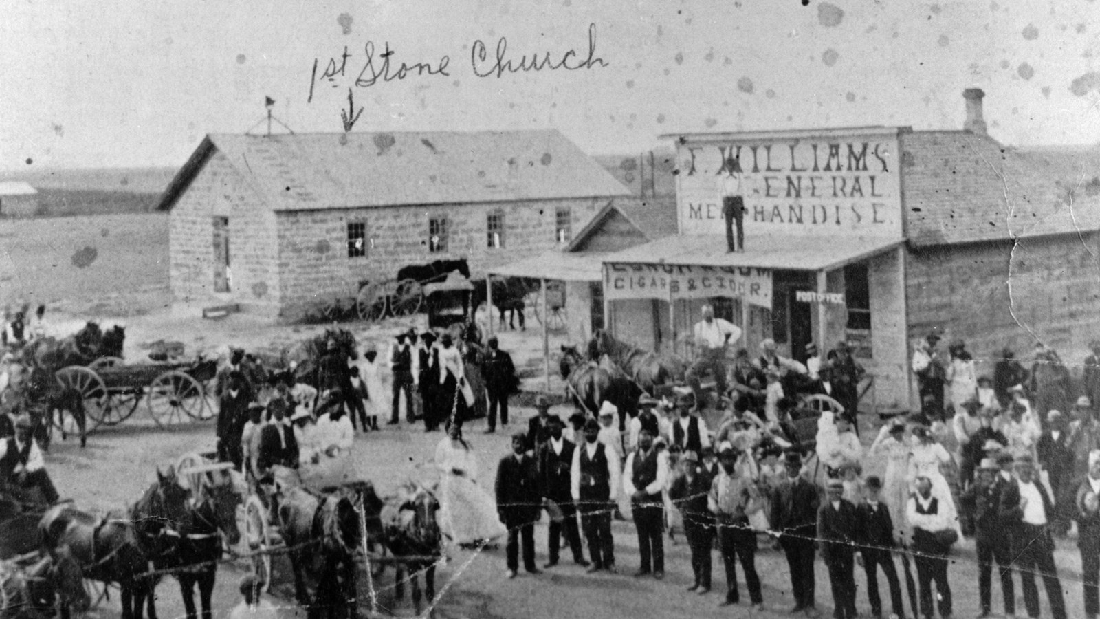 Black and white image of town street with many people gathered in front of general store.