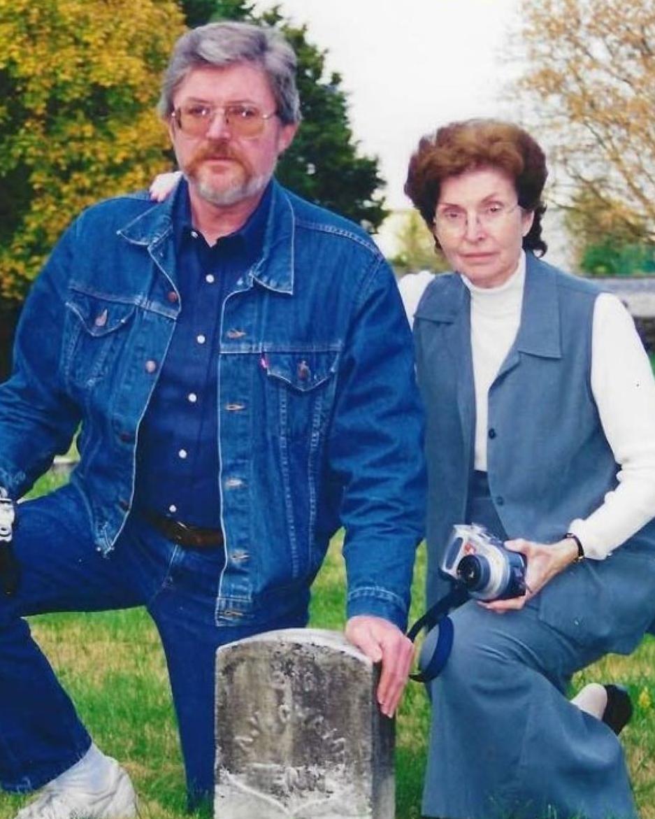 Man wearing all denim and woman wearing blue and white posed next to A.V. Champ's headstone. 