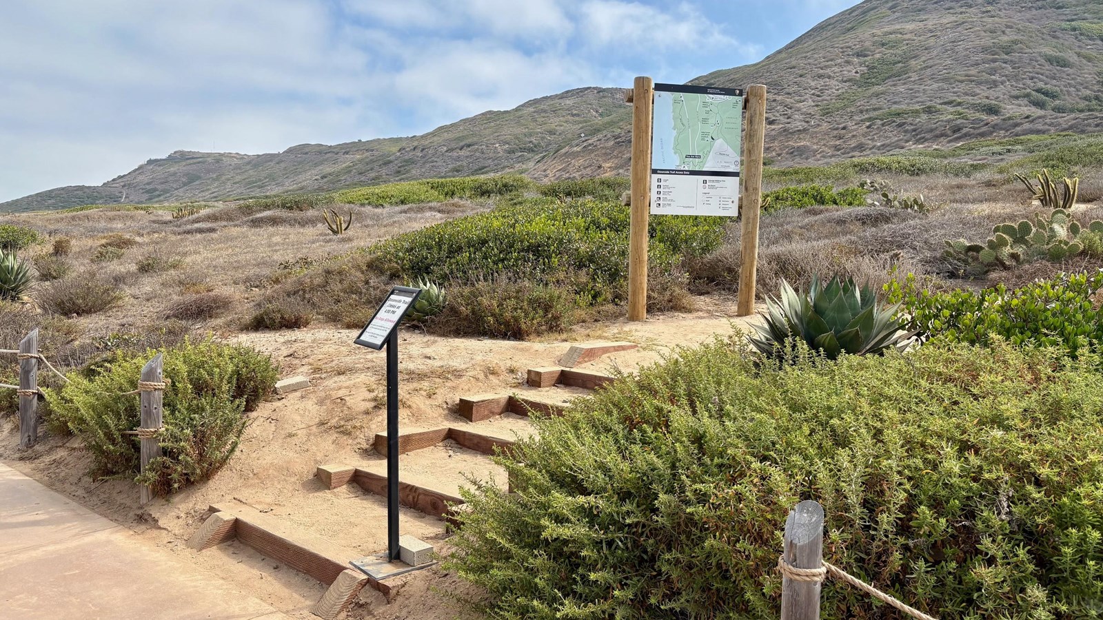 A trailhead sign adjacent to wooden steps with hills in the background.