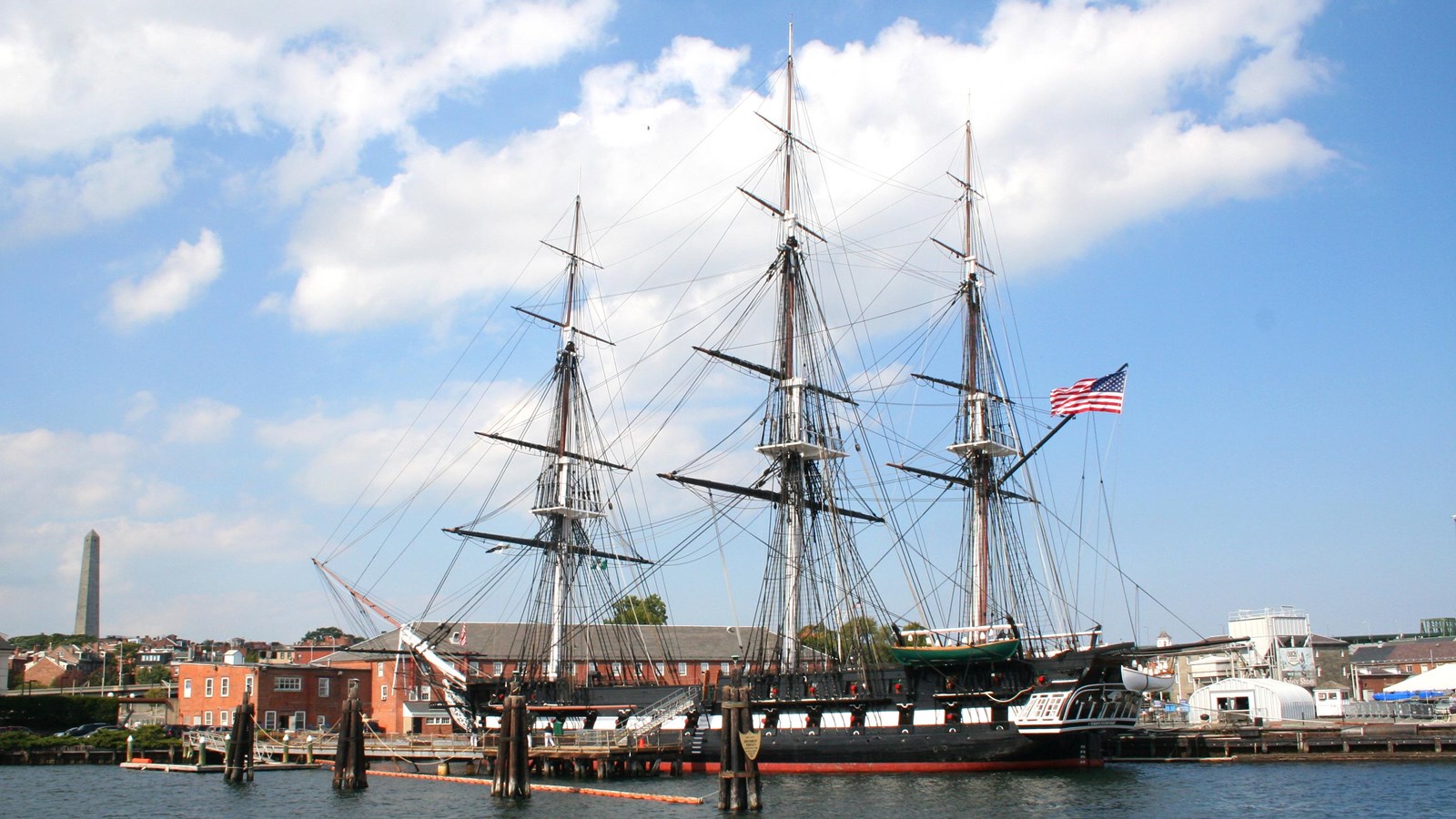Photograph of a three masted ship in water with blue sky and some clouds.