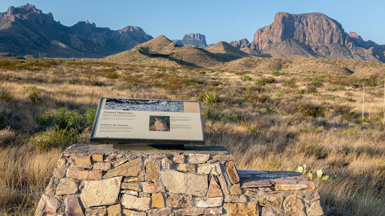 A wayside panel mounted on a stonework base with views of jagged mountains in the background