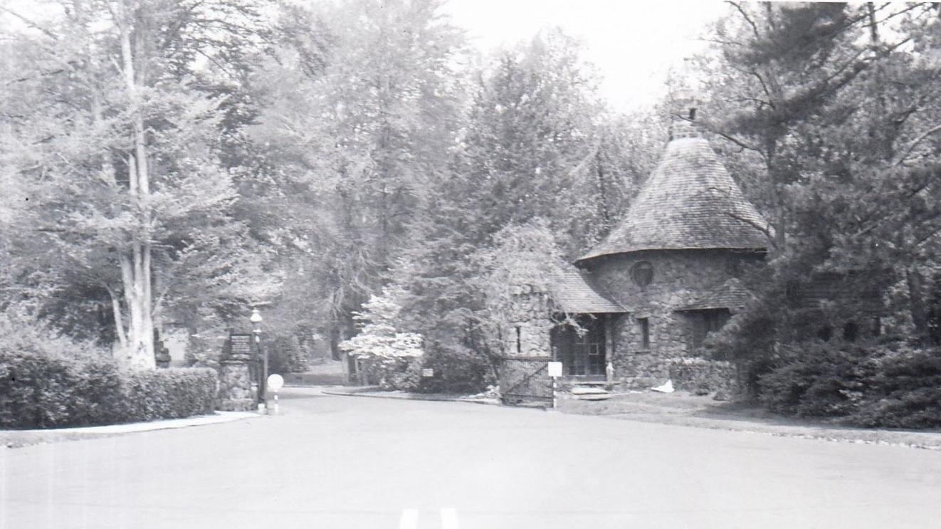 Black and white photo of small stone structure with road and gate surrounded by trees