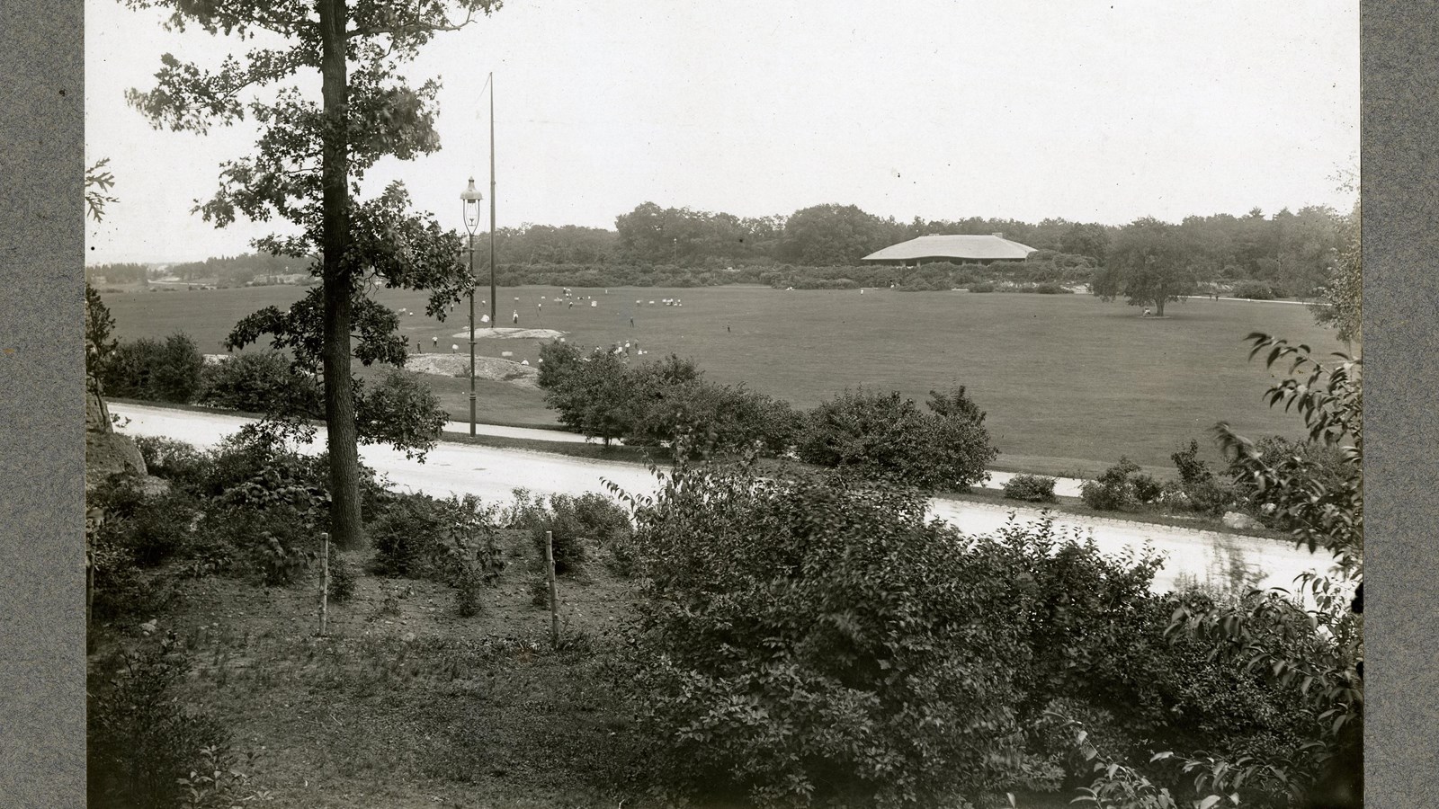 Black and white of flat grassy area with people on it with trees around all edges
