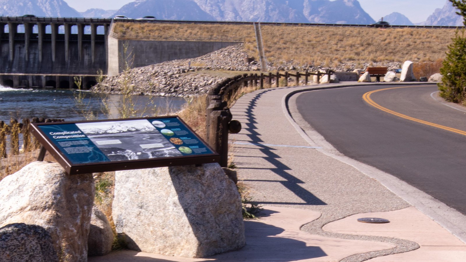 A panel with Jackson Lake Dam information by a concrete path with the dam in the background.