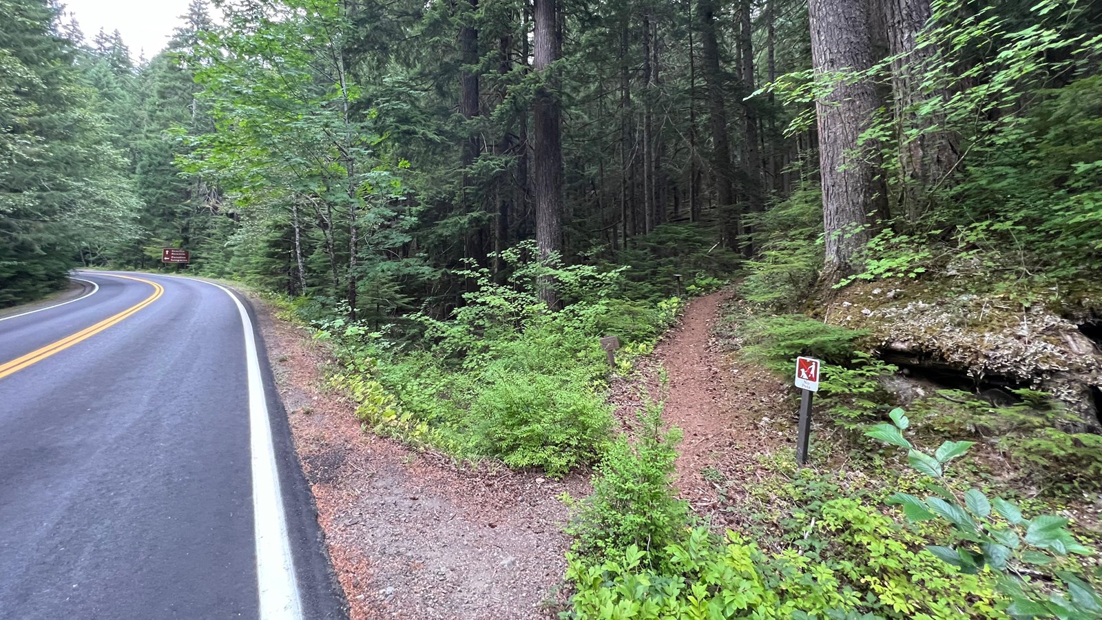 To the east side of the two-lane paved highway is a narrow, dirt trail surrounded by trees.