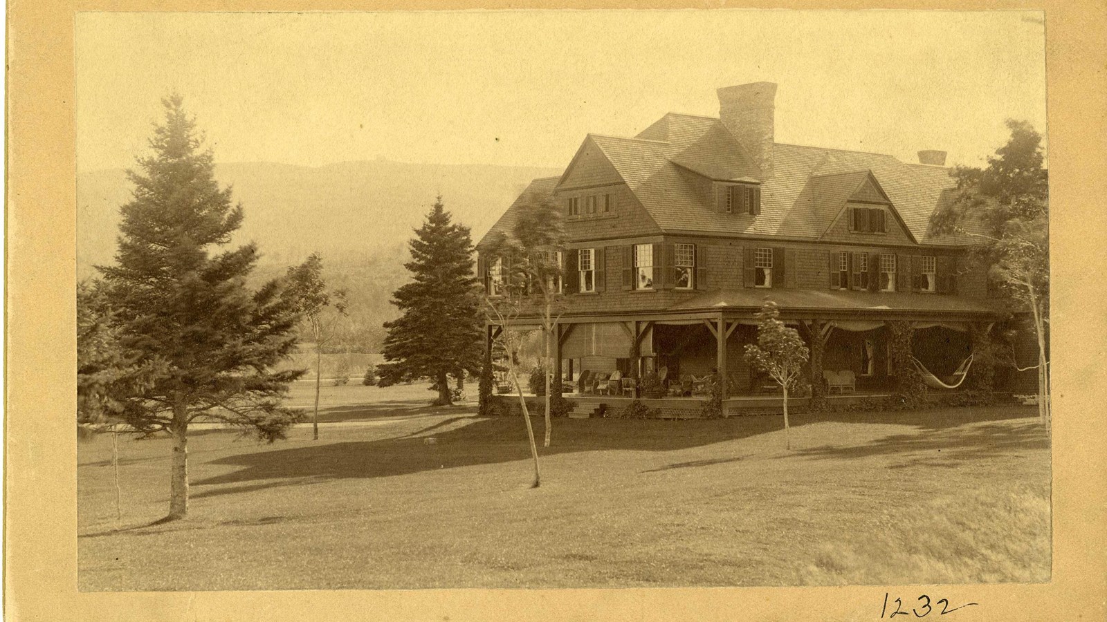 Black and white of large wooden home with flat grassy area around with trees spread out