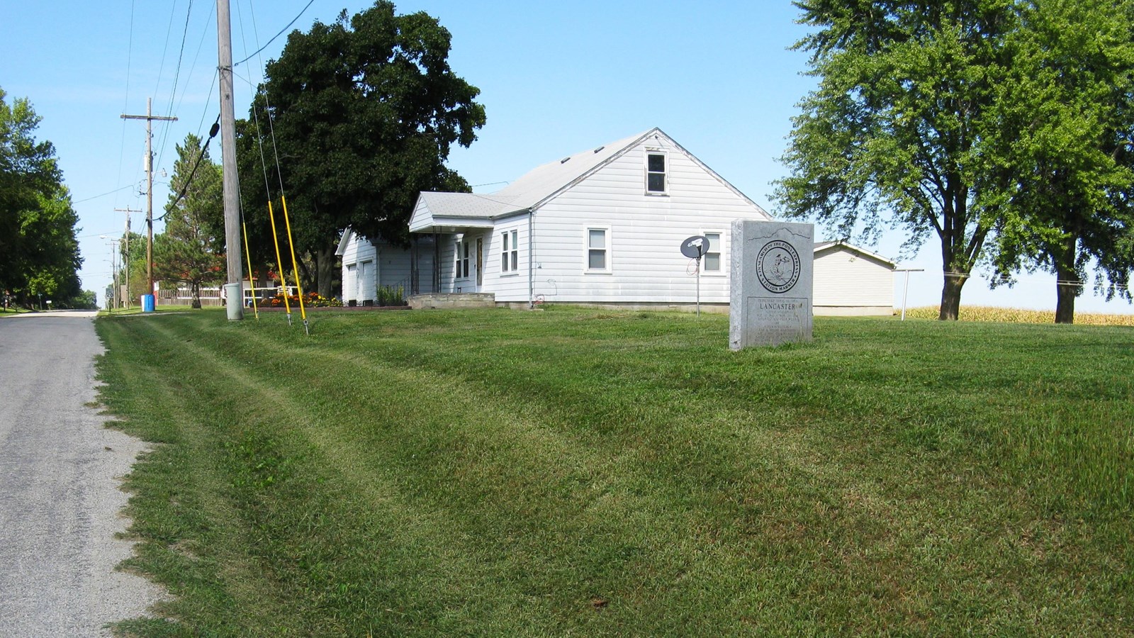 A gray monument stands on grass between a paved road and small trees. A white house in distance.