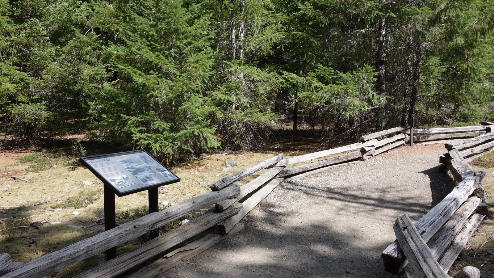 An exhibit panel stands along a gravel path bordered by a split-rail fence.  