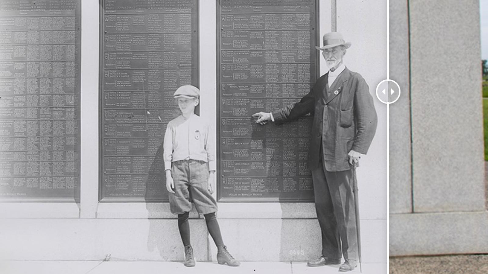 Black and white photo of an elderly man pointing to a bronze plaque. A young boy stands nearby. 