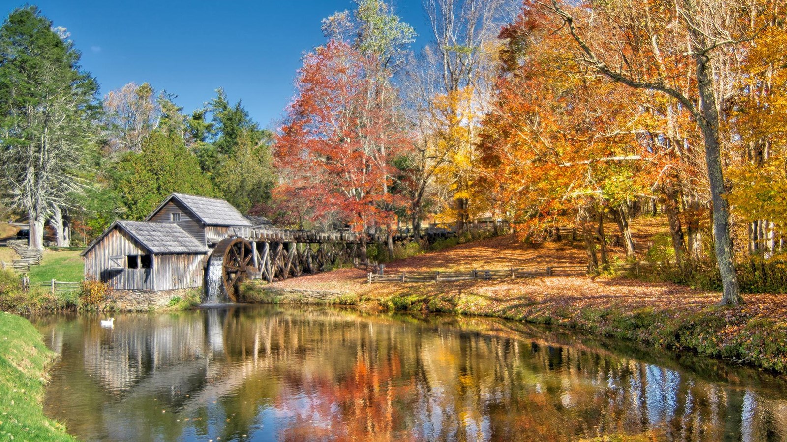 Autumn colors reflect in the water on a mill pond. On the far bank sits an old, weathered mill