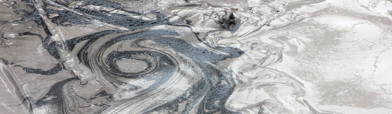 Swirls of black, gray, and white flow through a bubbling mudpot.