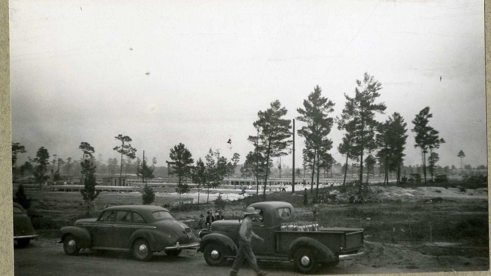 Black and white of dirt and grass area with many trees and cars along the side