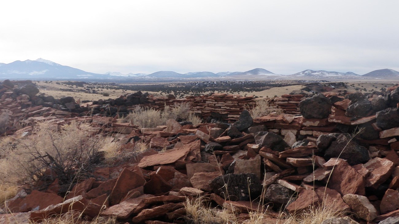 The remnants of a sandstone and basalt pueblo structure with volcanic hills in the background.