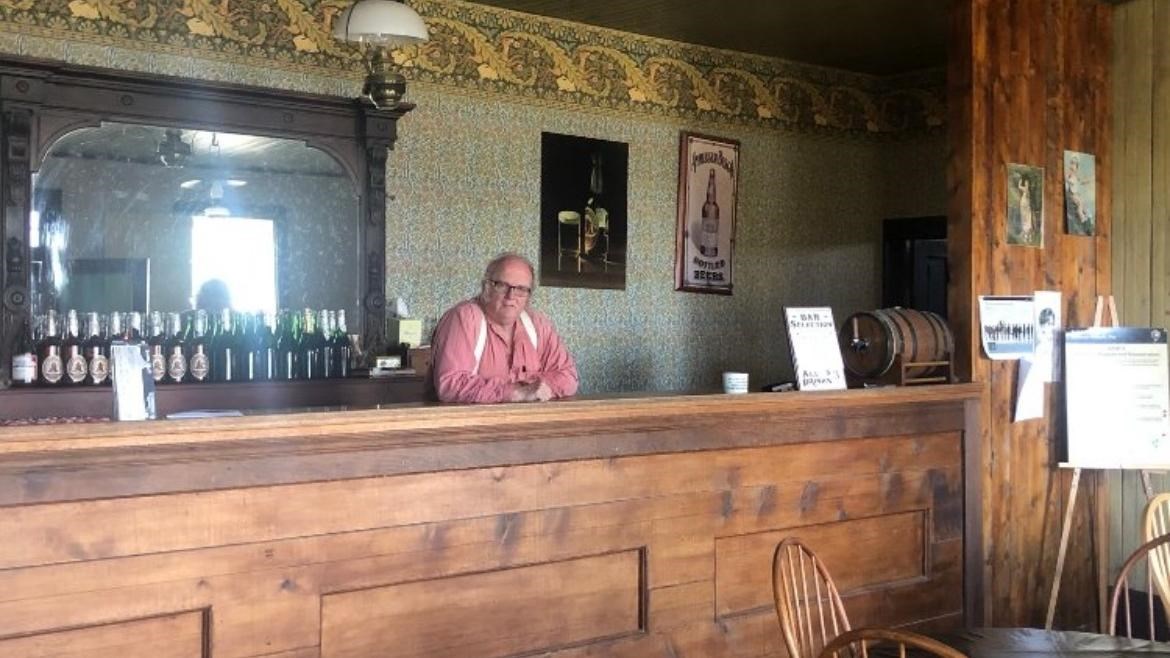 A man is standing behind a wooden bar with a large mirror right behind him.