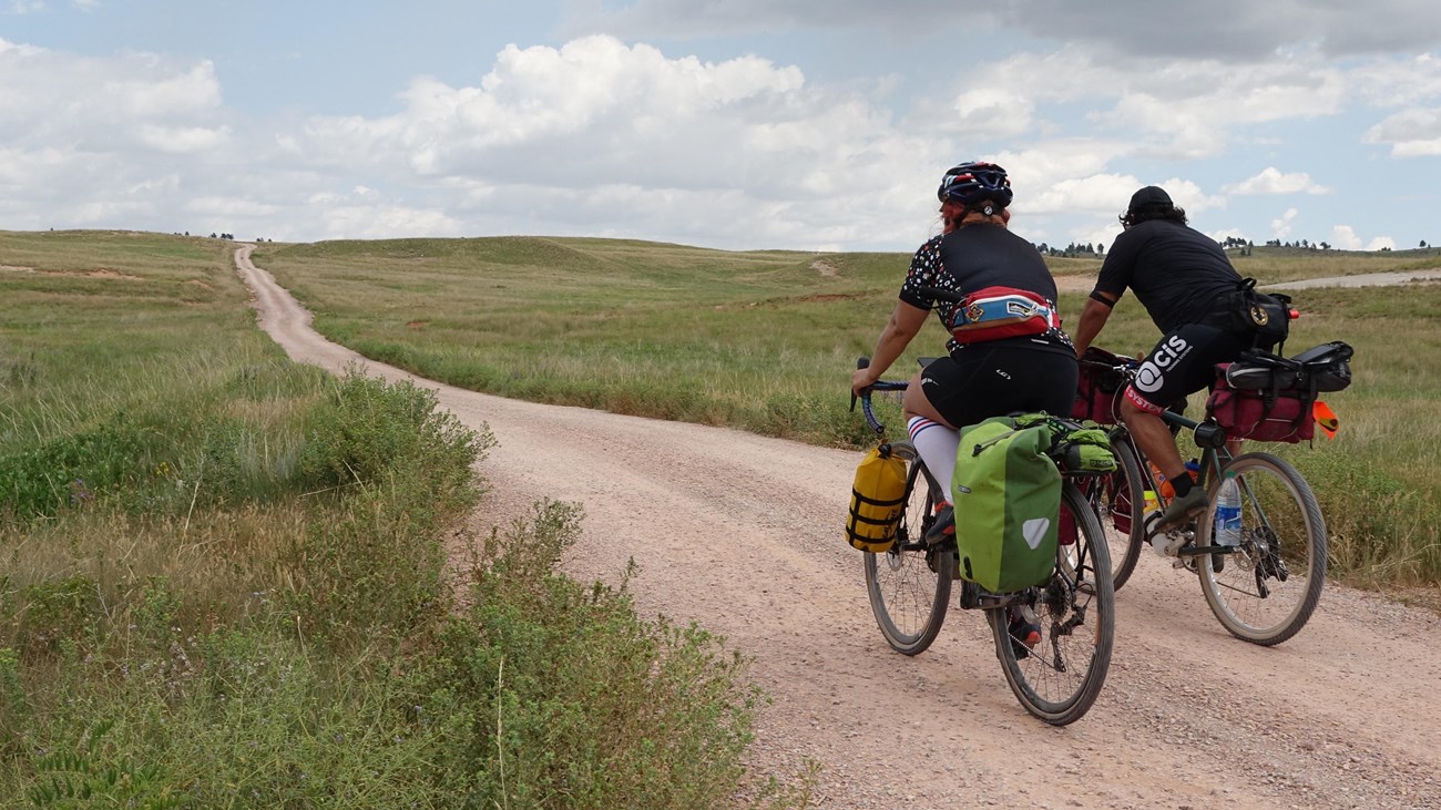 two people in biking gear riding bikes on a dirt road in the open prairie