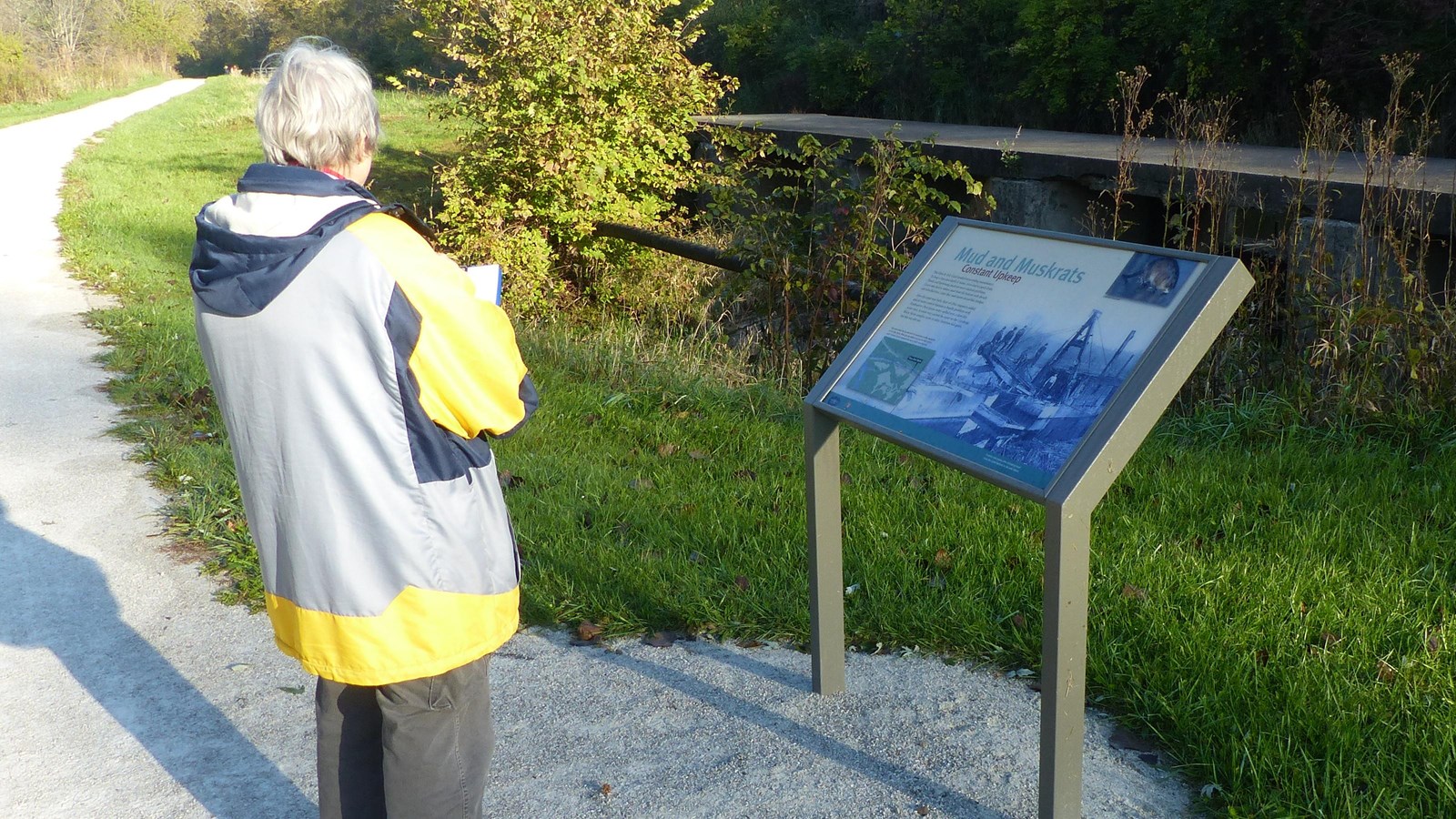 A grey-haired woman in a raincoat stands before a graphic panel along an unpaved trail.