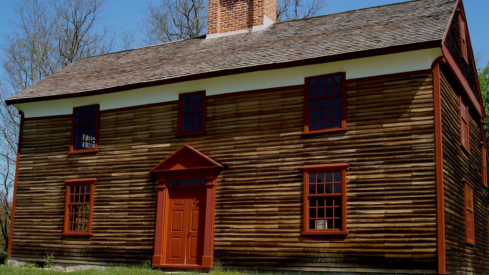 Two story wooden colonial house, unpainted wooden siding and a central chimney. 