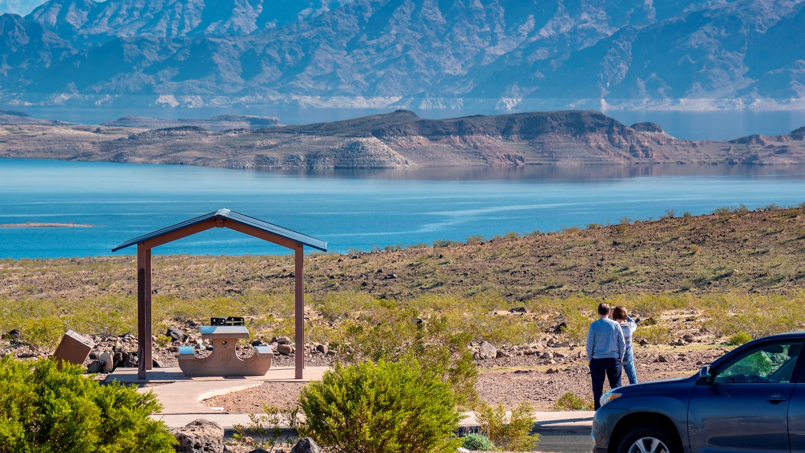 Two people walk on a sidewalk next to a parking lot looking out over a large lake.