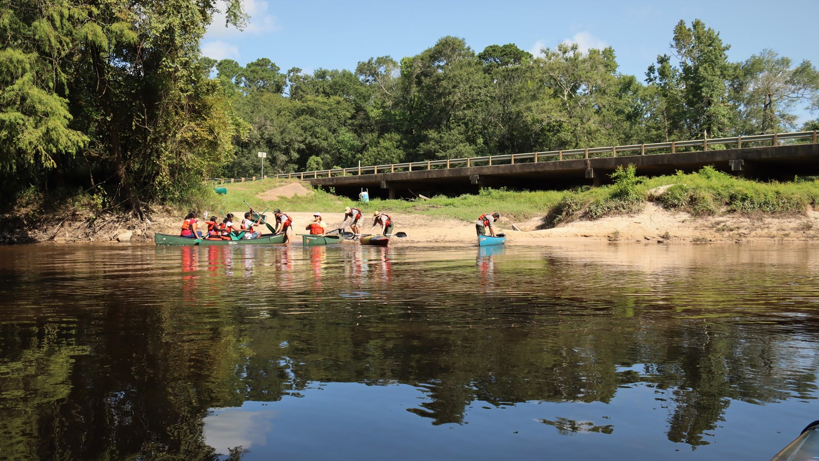 People launching several canoes onto a creek from a dirt boat ramp next to a highway