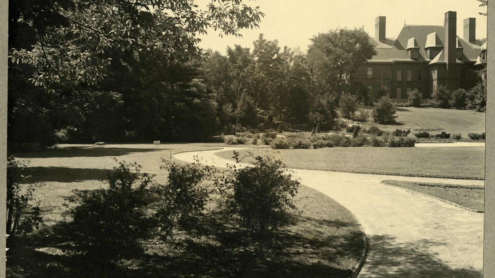 Black and white of curving paths around flat grassy area with trees on edges, leading to building