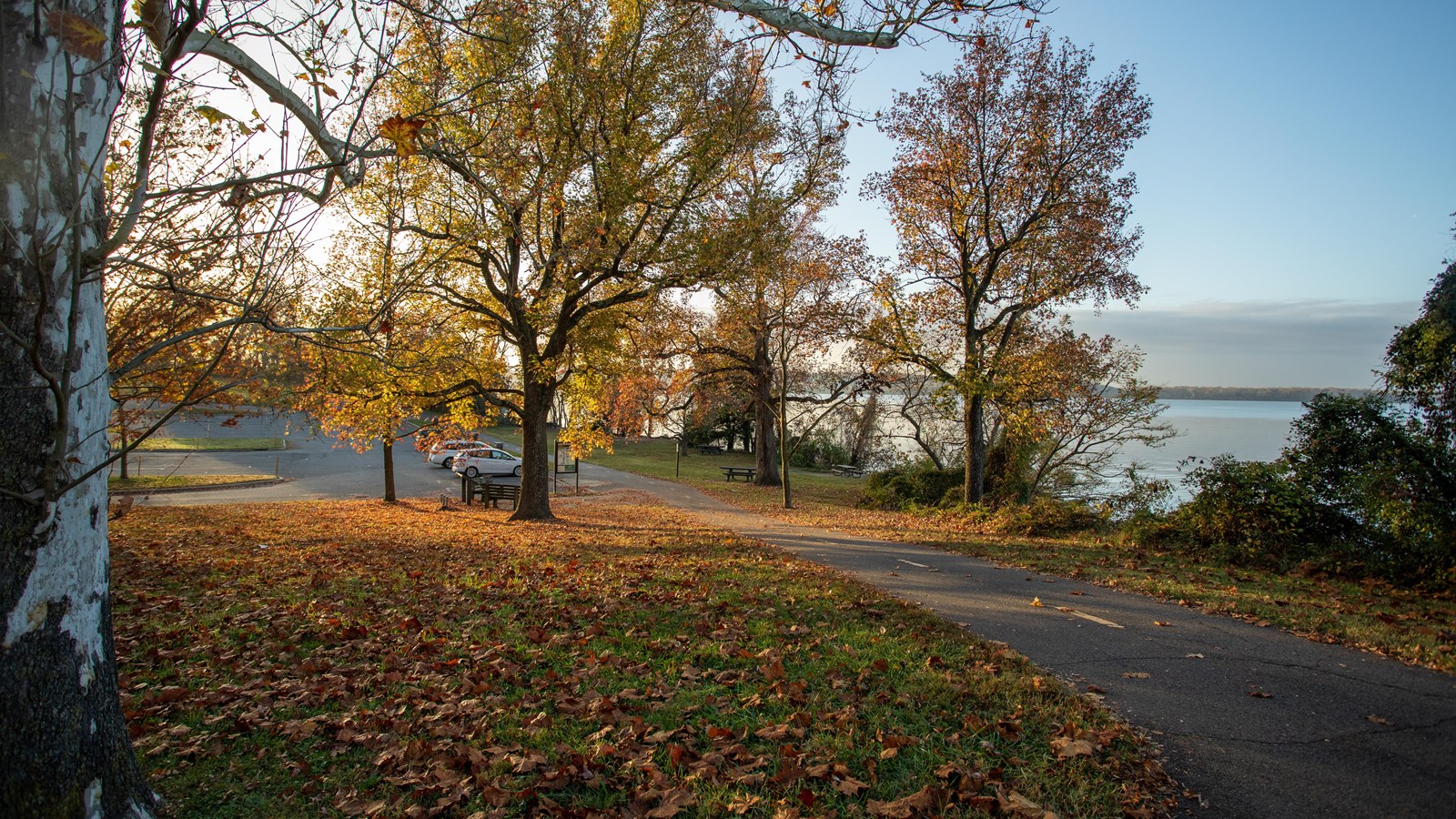 A pedestrian pathway is positioned next to a blue river of water. A parking lot is seen in the back.