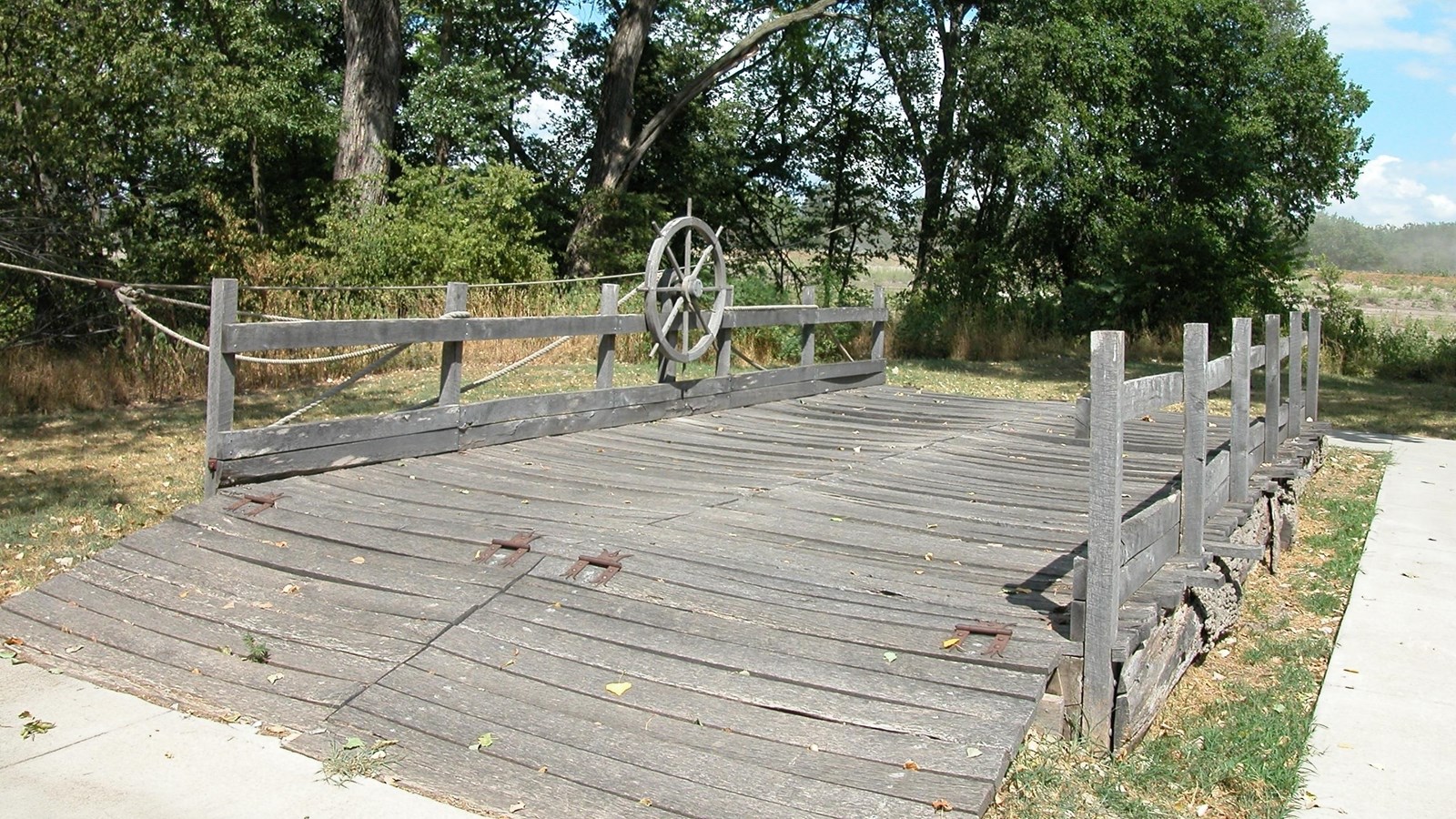 Wooden bridge surrounded by trees and a concrete path the the right.