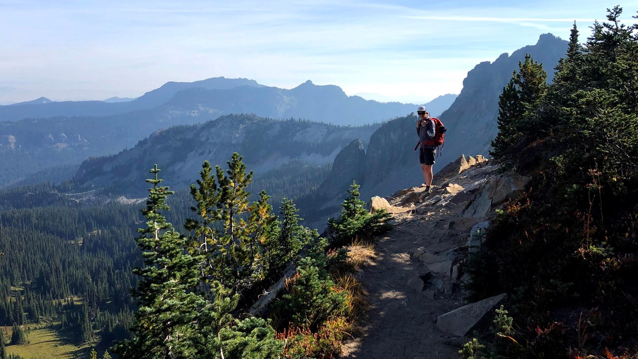 A backpacker on a rocky trail pauses to overlook a forested valley and mountain ridges. 