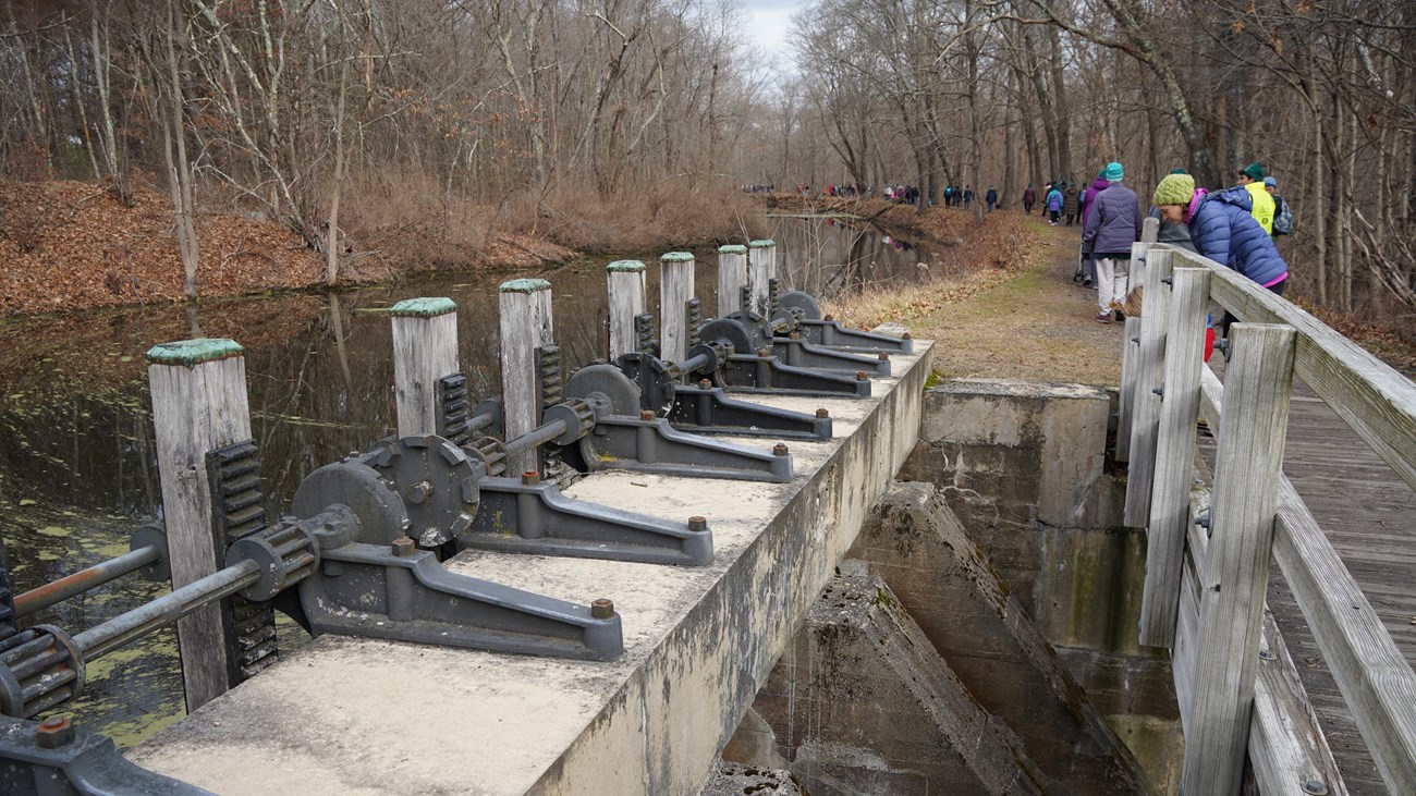 Group of people walking down path with industrial mechanisms visible on the left side of image