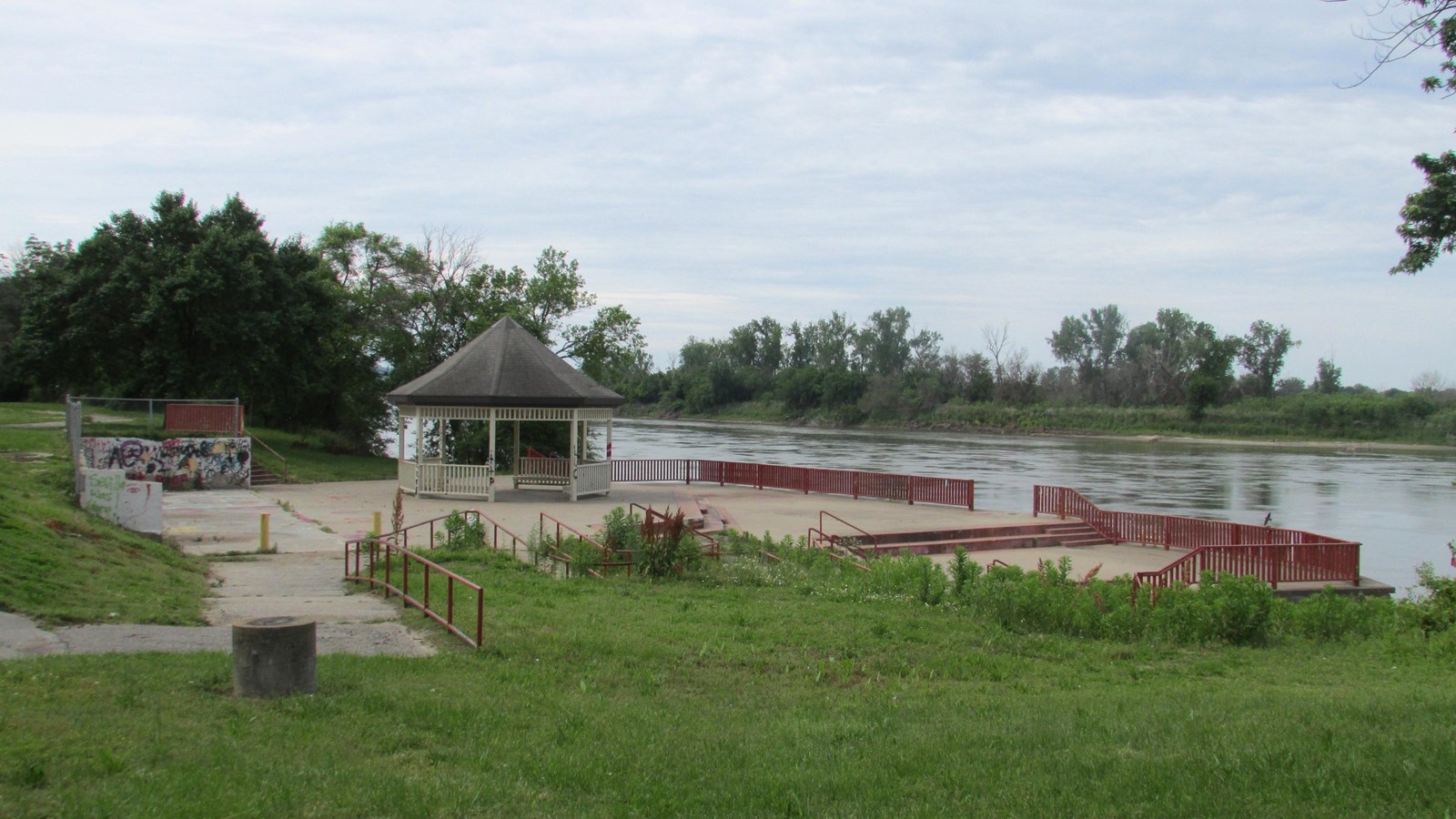 Beyond a patch of green grass, a peak-roofed gazebo stands on a concrete platform at edge of river.