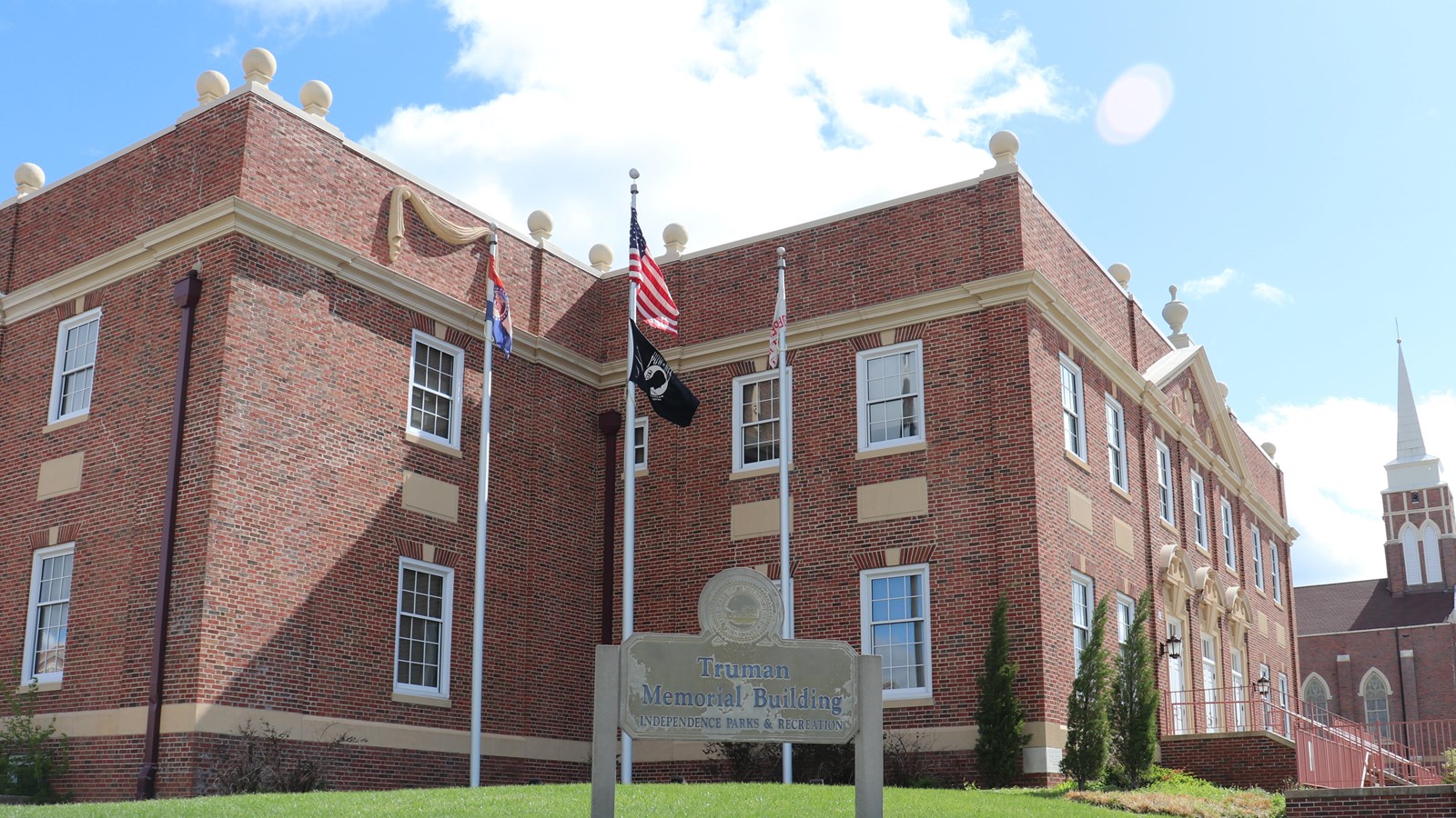 In the foreground, 3 flags on a flag pole. In the background, a 2 story brick building.