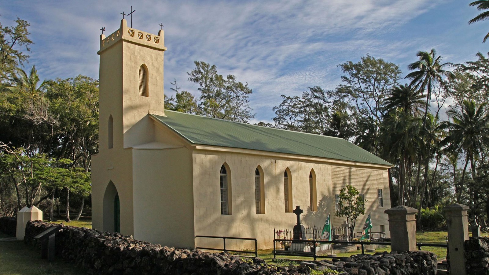 A cream white building with a steeple and a low rock fence in front. 