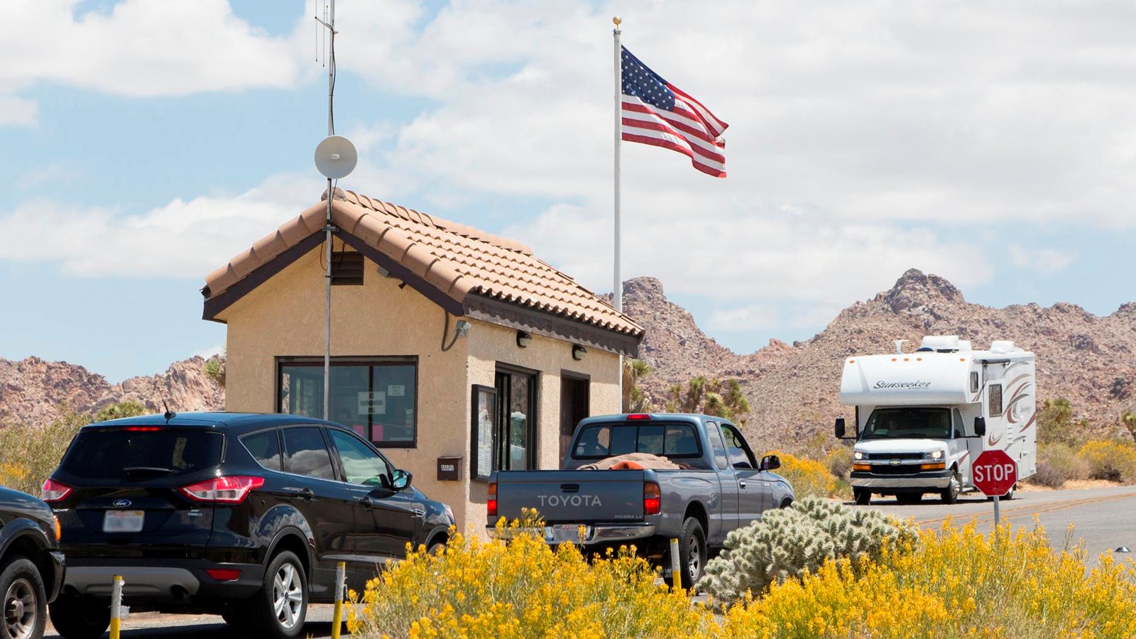 A line of cars, a small tan building with large windows, and a flag pole flying an American flag. 