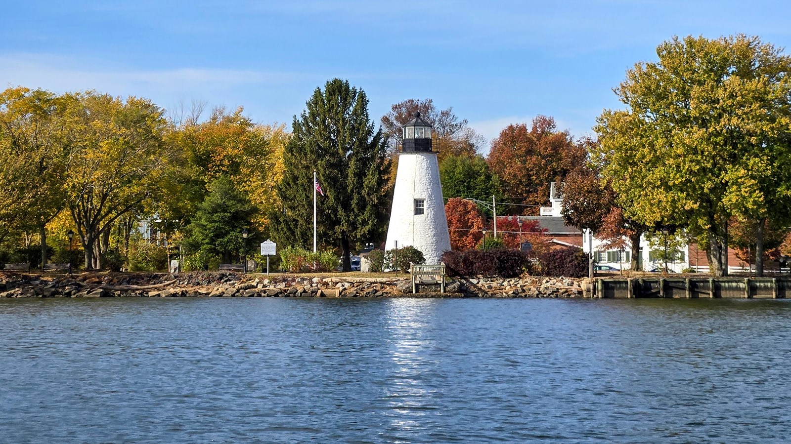A lighthouse with white body and black top with autumn trees and a body of water in the foreground.
