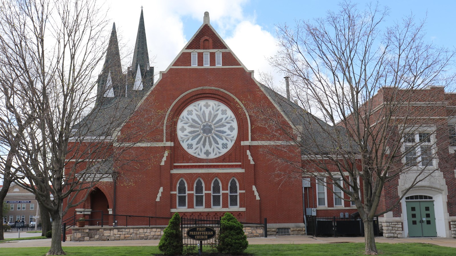 A large brick church with triangular roof, decorative circular window, and two steeples.