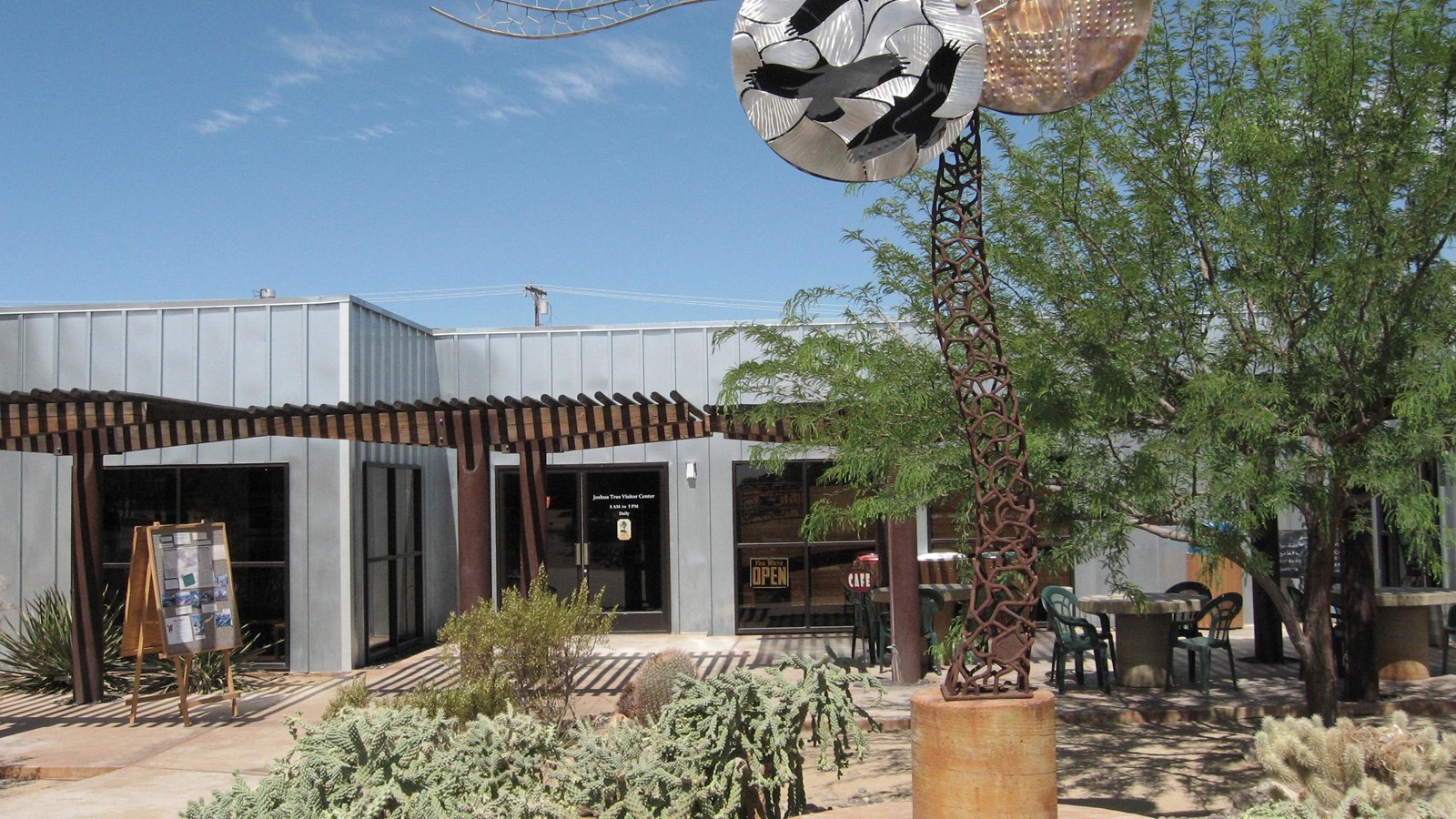 A large gray building with brown shade structures. In front is a large metal art piece and cacti.