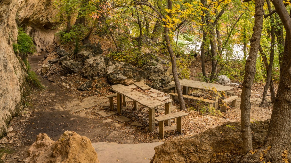 Stone tables and benches are located near a cliff with rock overhangs and alcoves