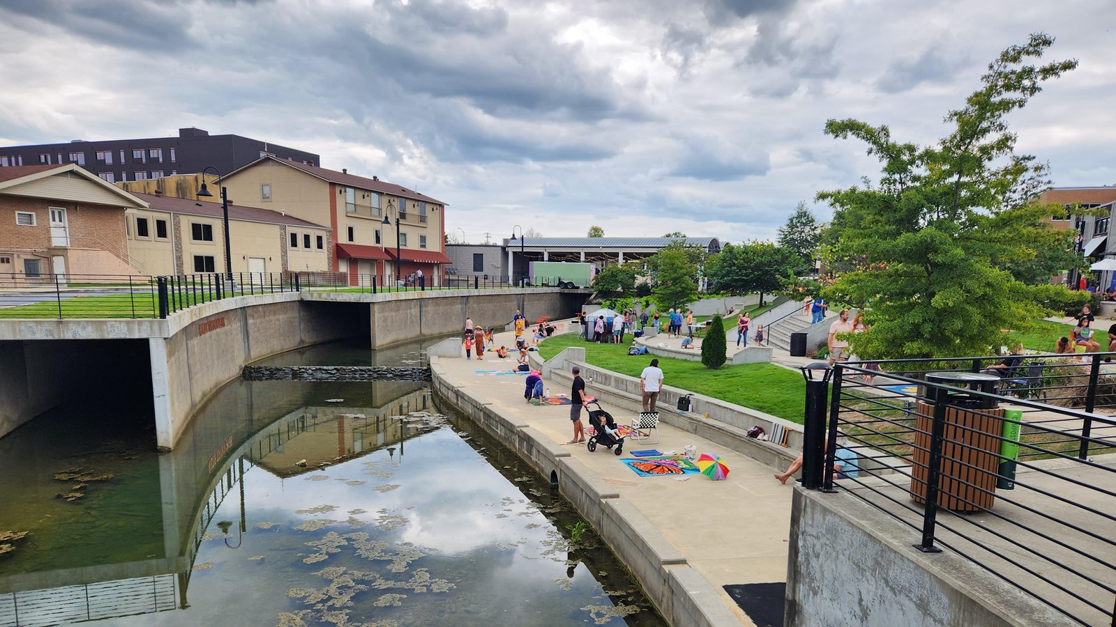 A sidewalk runs alongside a canal. There is a strip of grass and trees.