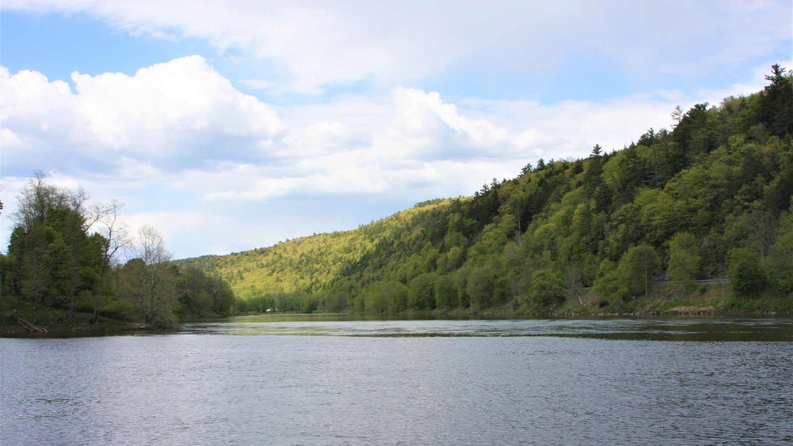 Pale blue river reflects cornflower sky with clouds. Green tree-covered hills in background.