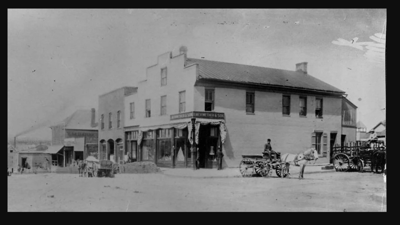 Historic image of a two story wooden buildings lining a wide main street with people on wagons.