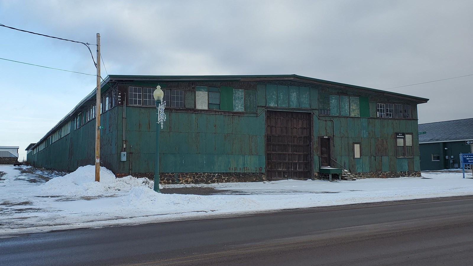 Green single story corrugated building with windows on top and a double door main entrance. 