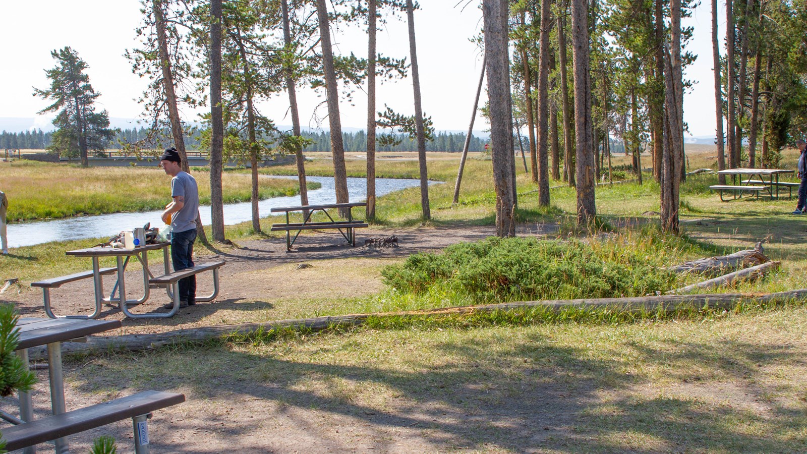 A person prepares a meal at a picnic table near a stream.