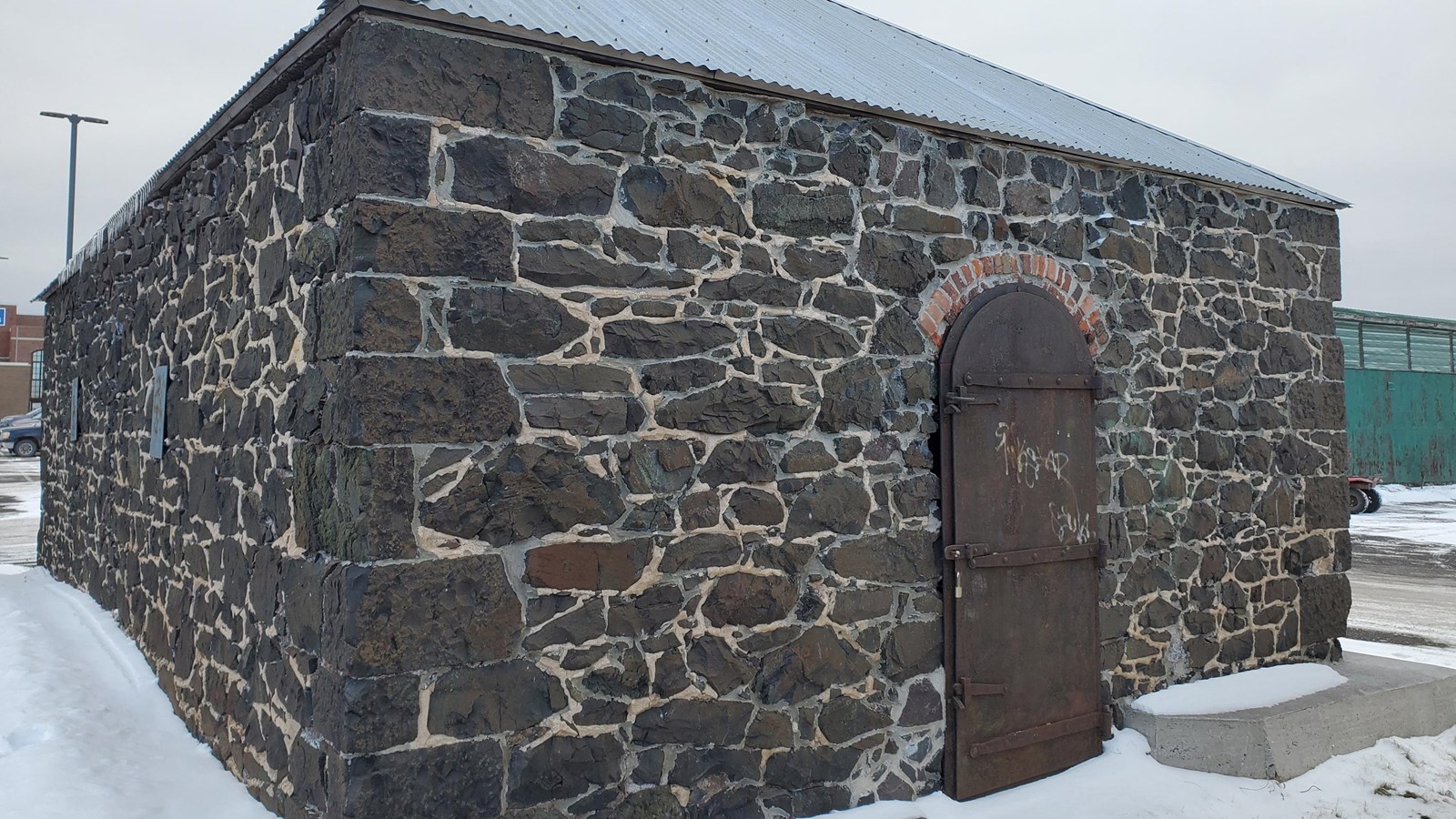 A poor rock building , a heavy metal door with brick arch above, and a corrugated metal roof. 