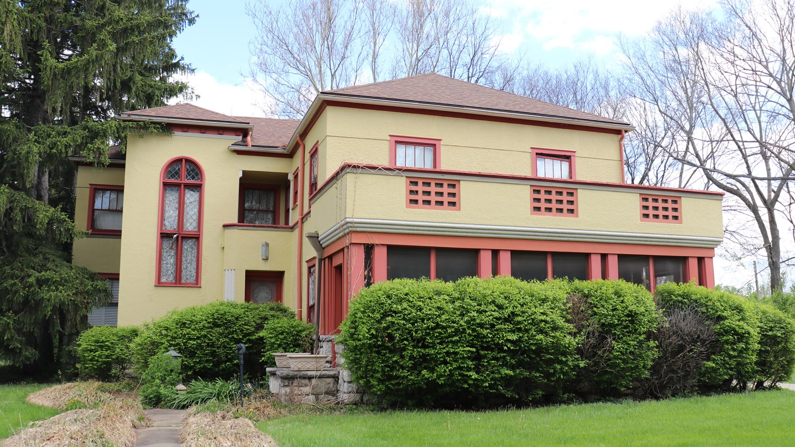 A yellow and red two story home with a screened in front porch and bushes surrounding the home.  