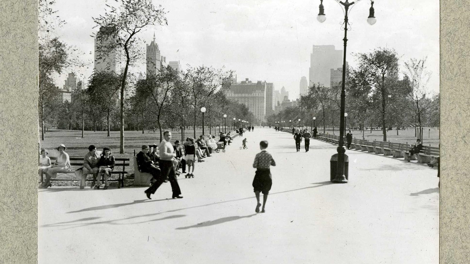 Black and white of large path lined with benches and people walking on it and grass on other side. 