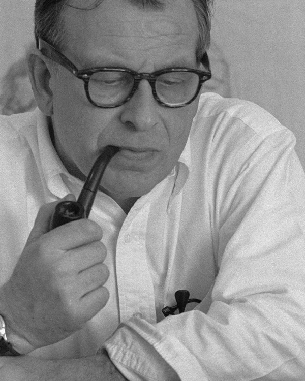 Man wearing white shirt and smoking a pipe at a desk. 