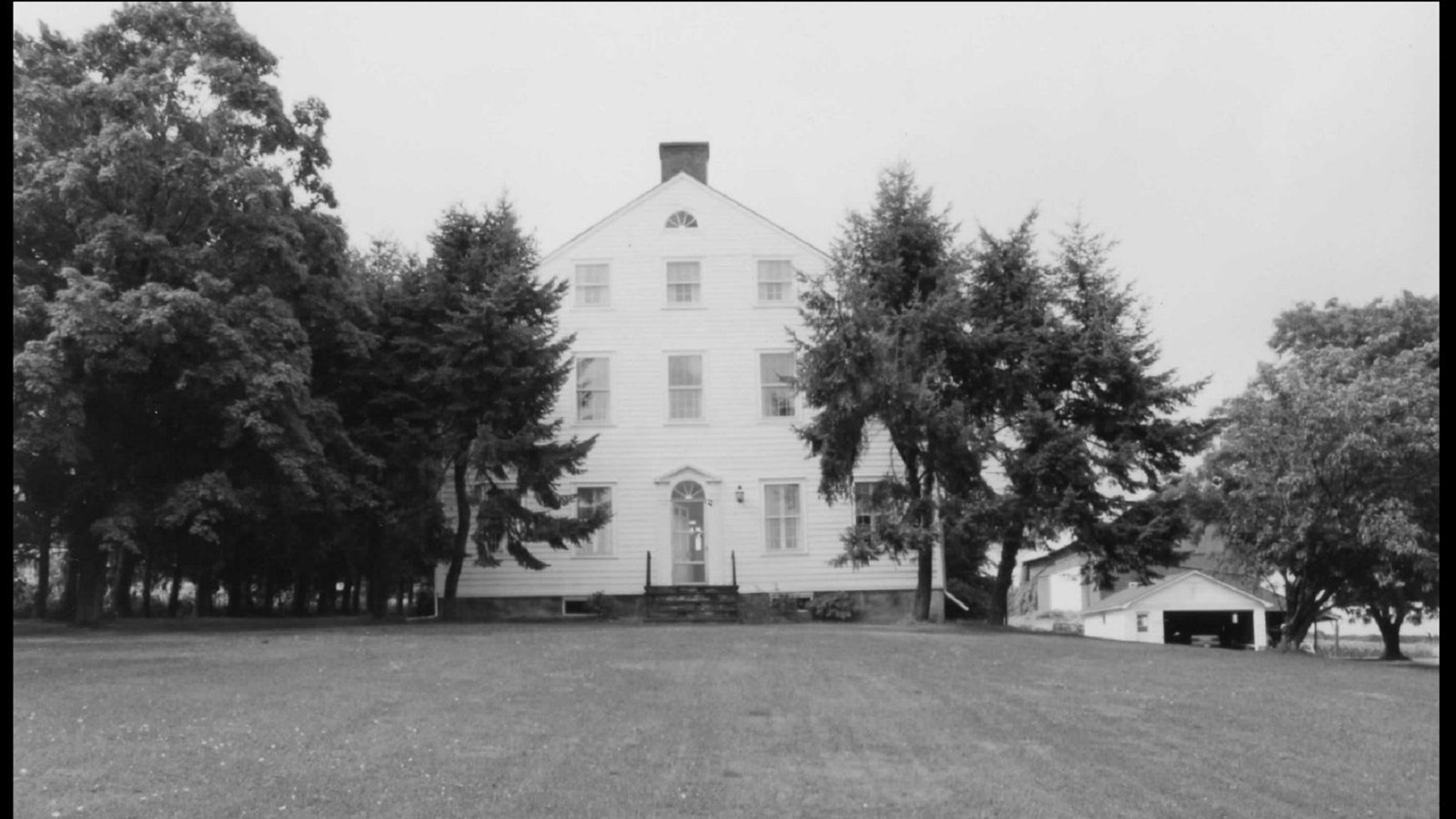 Black and white image of a white framed house framed by trees, with a grass lawn in foreground.