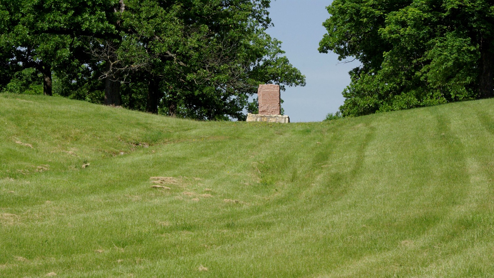 A stone monument stands in the foreground surrounded by grass and lush trees.