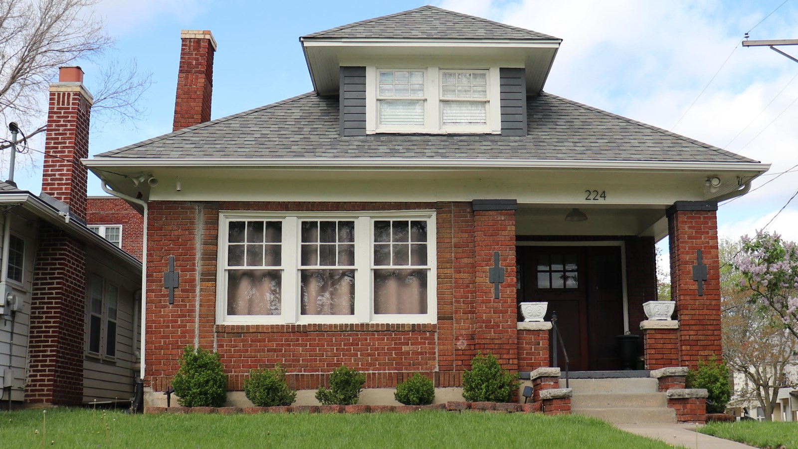 A one and a half story brick bungalow in background and green grass and bushes in the foreground.