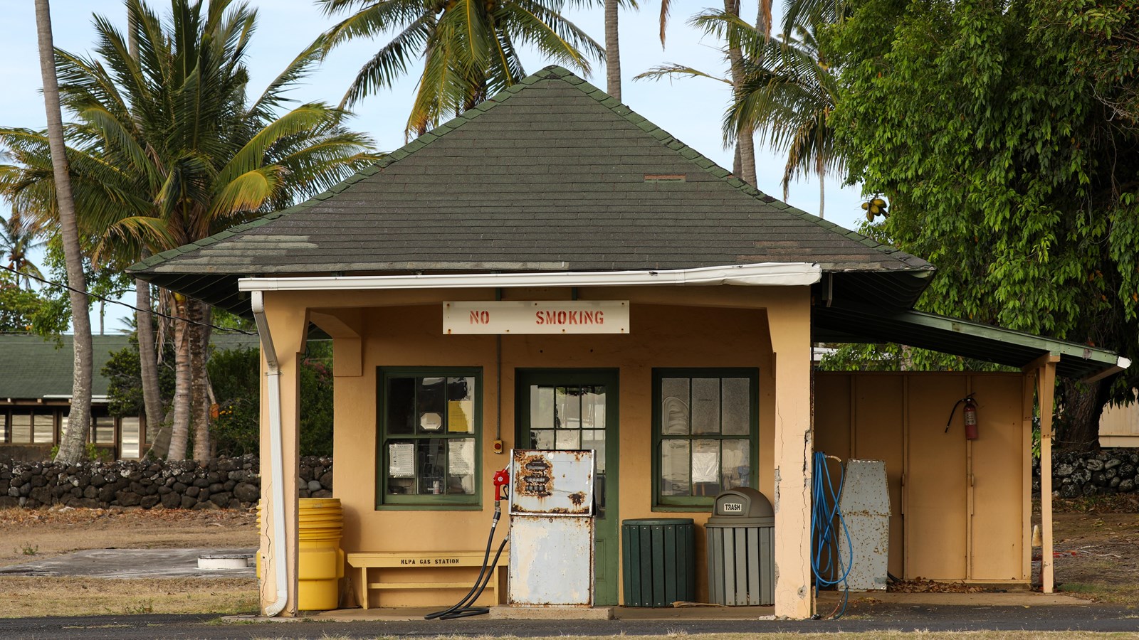 A tan building with a black shingled roof. In the front is a No Smoking sign and a gas pump.