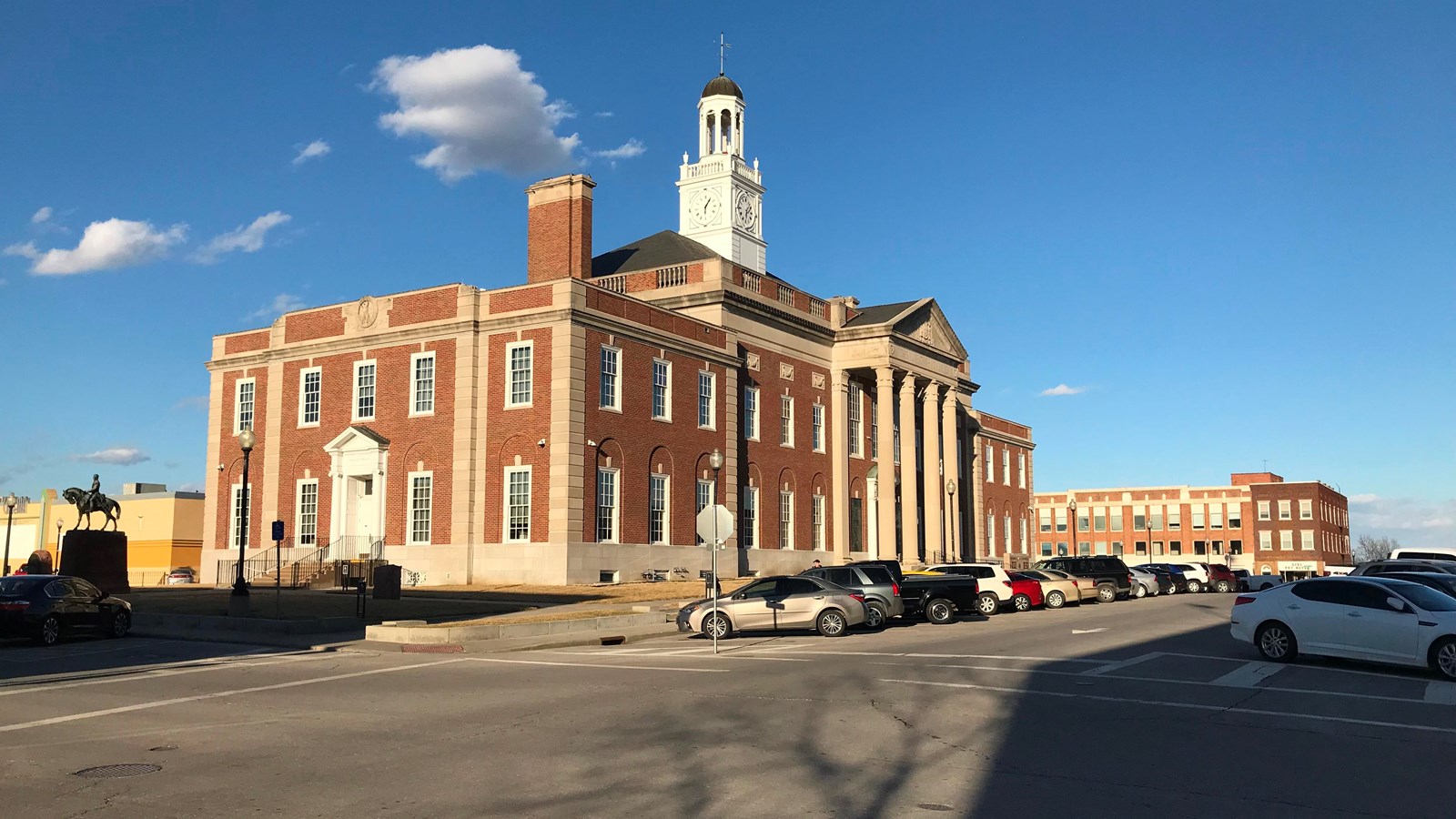 A 2-story red brick courthouse stands tall in the center with parking lot and other buildings around